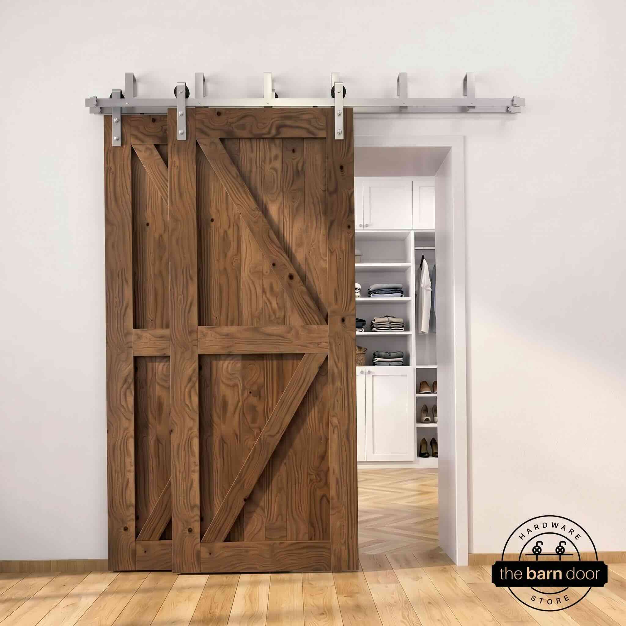 Brushed nickel double bypass barn door kit with two dark walnut K-brace doors mounted on a dual-rail track, partially open to reveal a white built-in closet with folded clothing, hanging garments, and shoe shelving, set against a white wall with light herringbone and oak plank flooring.