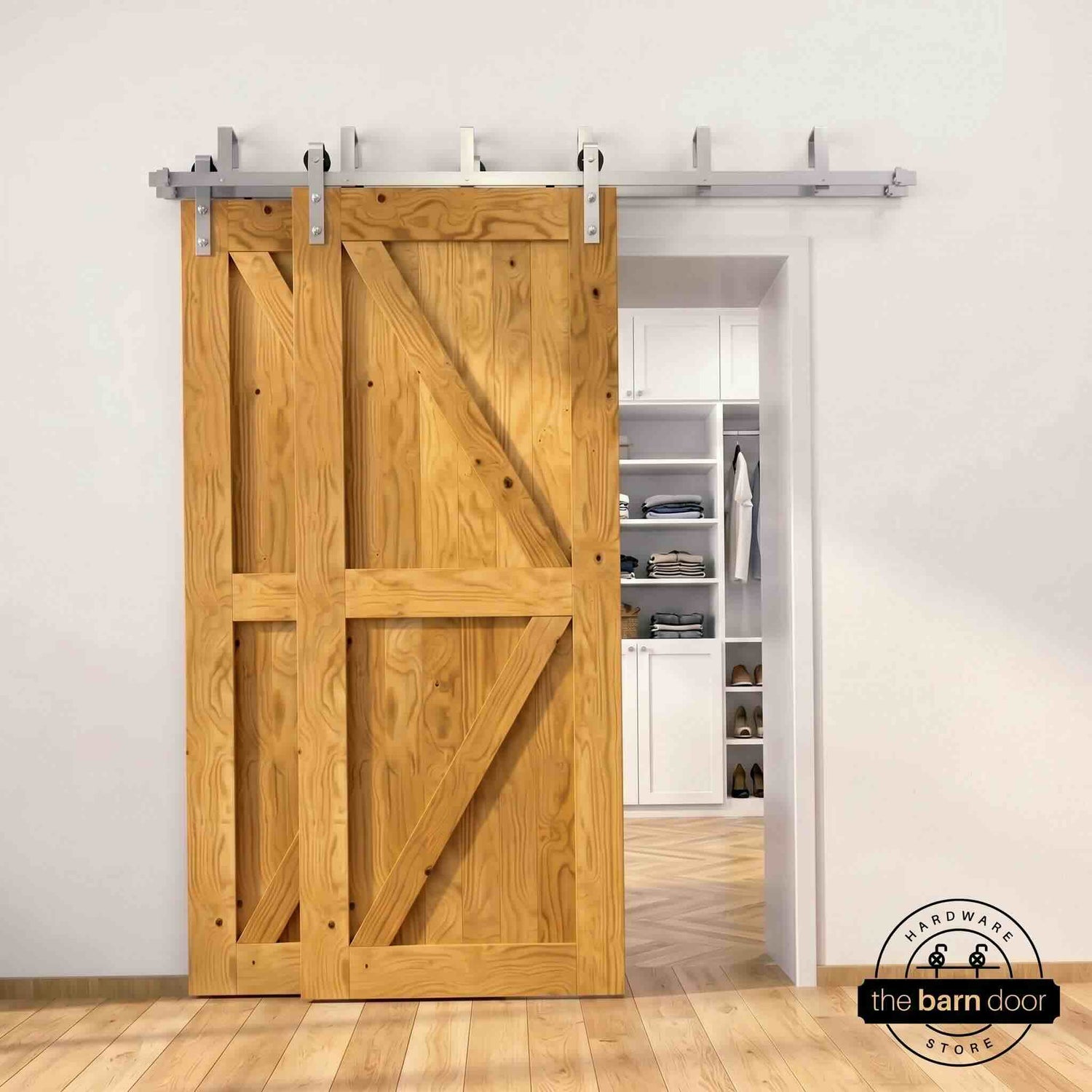 Brushed nickel double bypass barn door kit with two early american K-brace doors mounted on a dual-rail track, partially open to reveal a white built-in closet with folded clothing, hanging garments, and shoe shelving, set against a white wall with light herringbone and oak plank flooring.