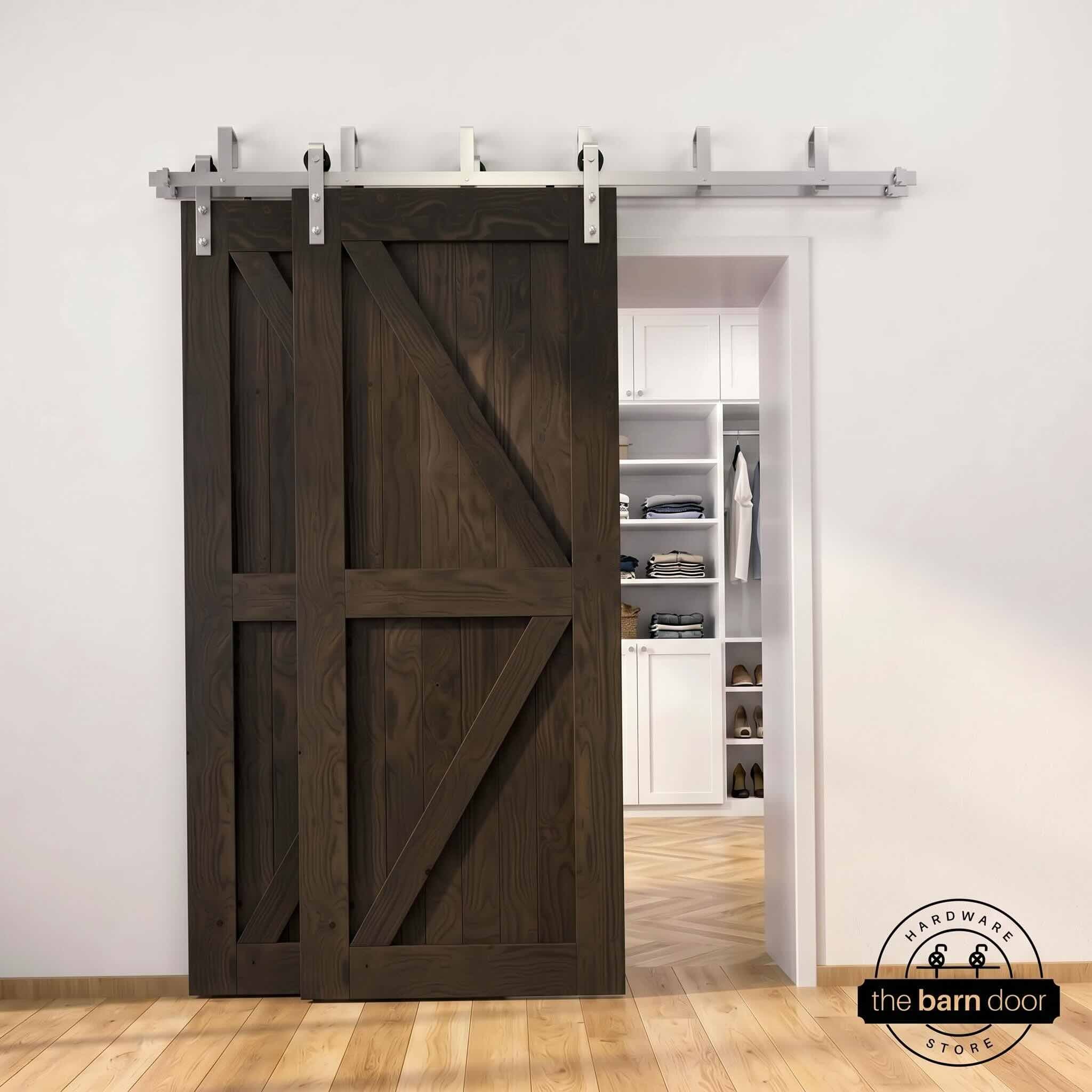 Brushed nickel double bypass barn door kit with two ebony K-brace doors mounted on a dual-rail track, partially open to reveal a white built-in closet with folded clothing, hanging garments, and shoe shelving, set against a white wall with light herringbone and oak plank flooring.