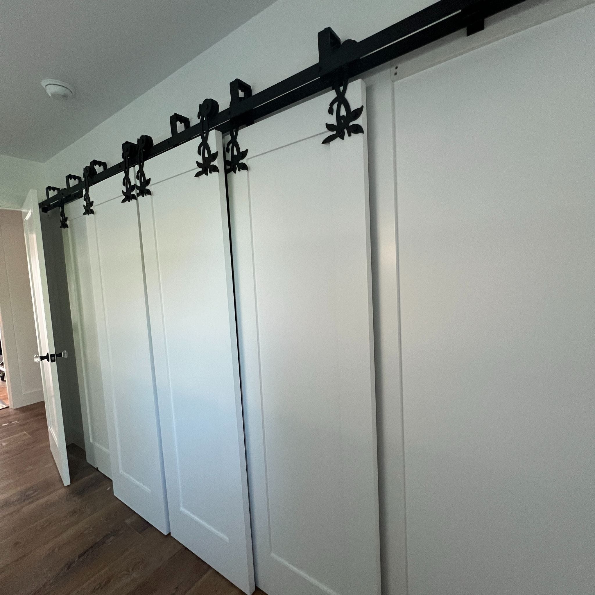 A wide-angle view of multiple white sliding bypass closet doors mounted on a continuous black double-track system featuring several ornate floral hangers.
