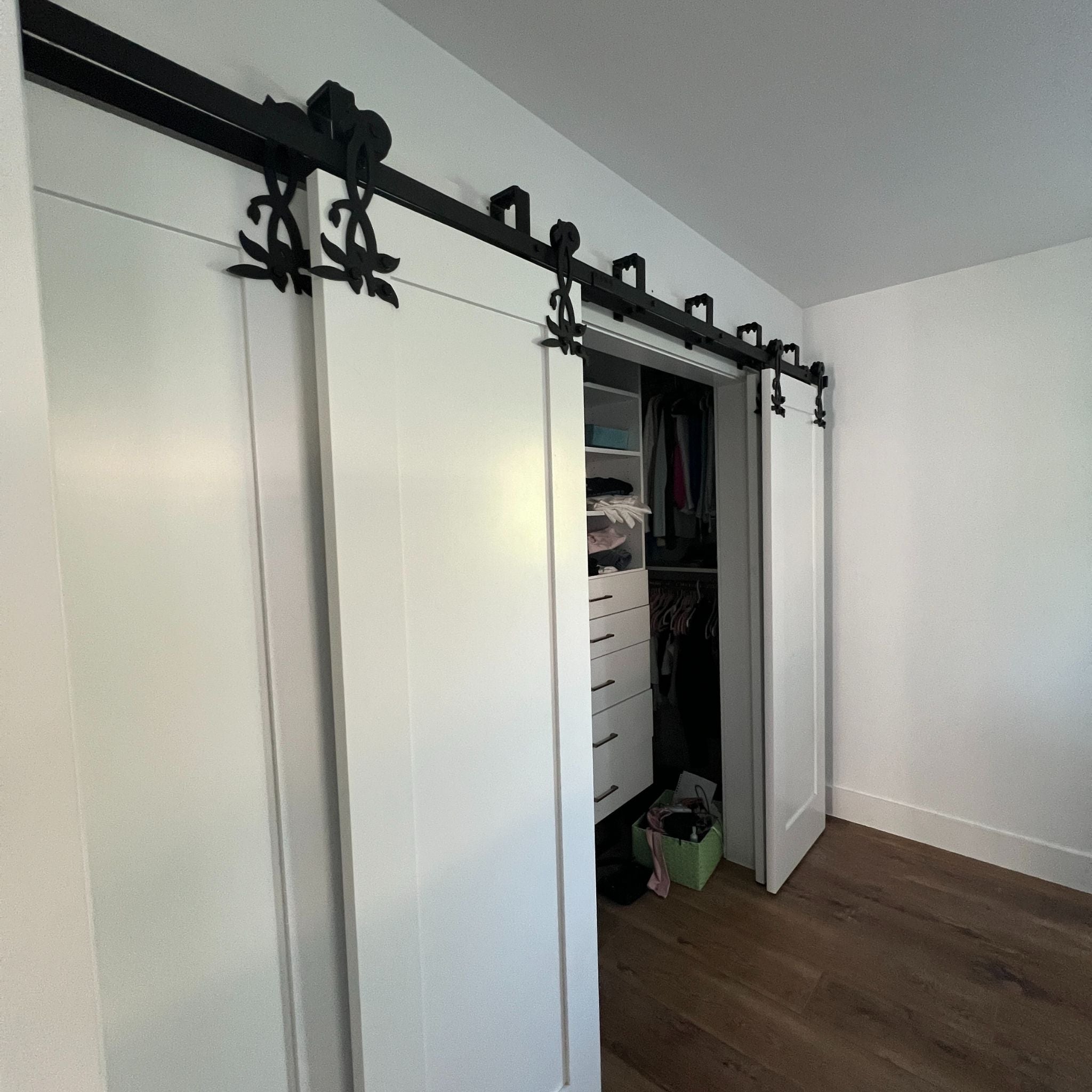 White bypass sliding closet doors with decorative black floral hardware, partially open to reveal an organized white walk-in closet with shelves and drawers.