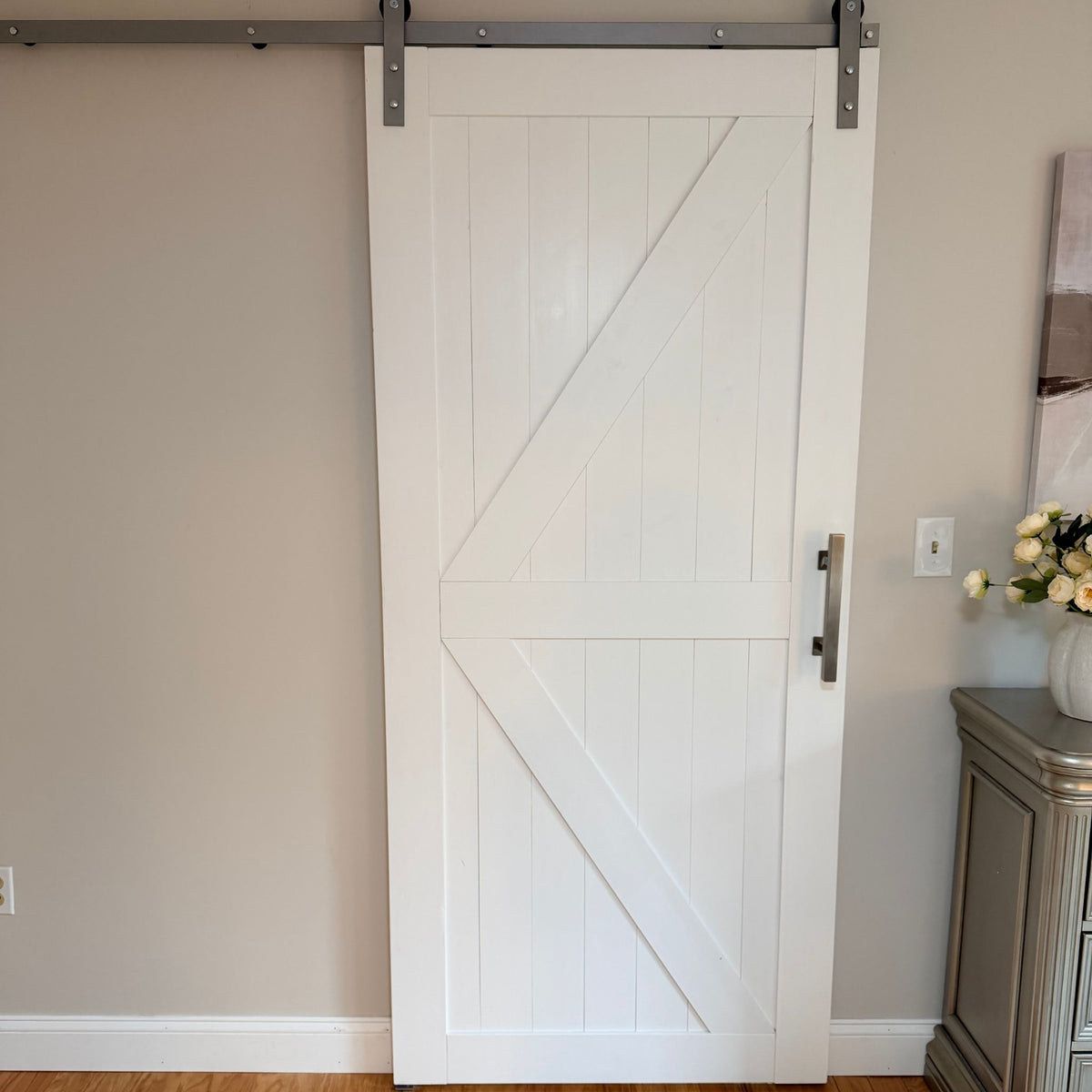 A white sliding barn-style door with diagonal X-brace panels and a vertical metal handle, installed on a metal rail against beige walls in an interior room.
