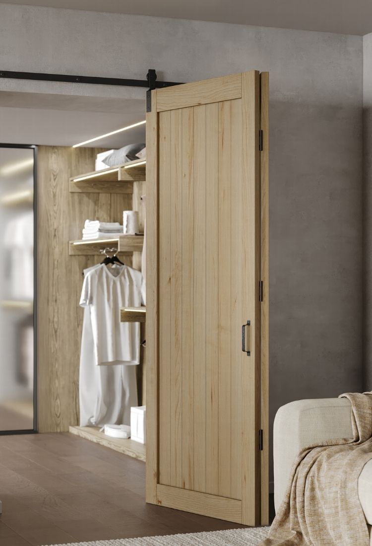 A panoramic view of a light wood bifold barn door system on a black track, open to reveal a lighted closet interior with wooden shelving.