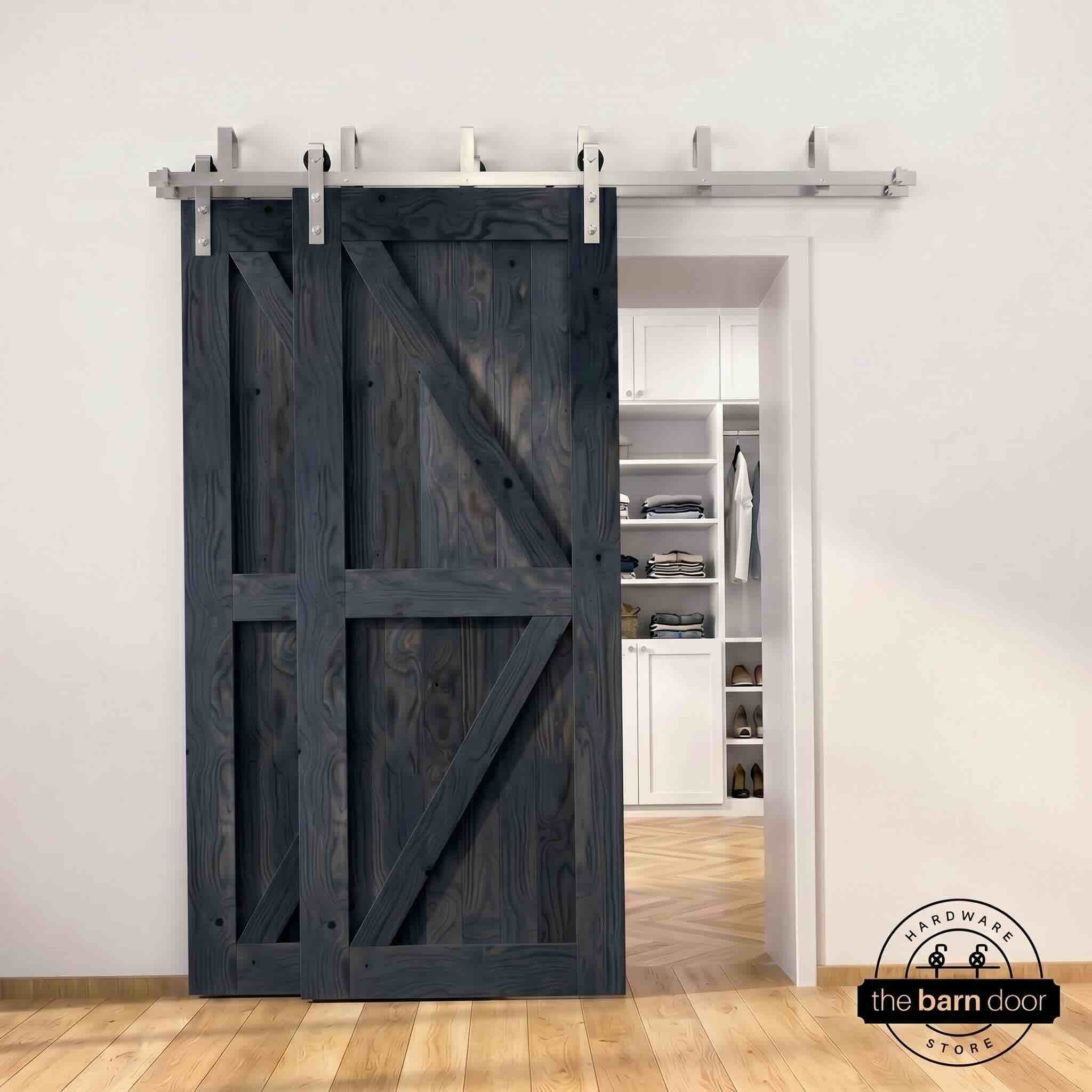 Brushed nickel double bypass barn door kit with two navy K-brace doors mounted on a dual-rail track, partially open to reveal a white built-in closet with folded clothing, hanging garments, and shoe shelving, set against a white wall with light herringbone and oak plank flooring.
