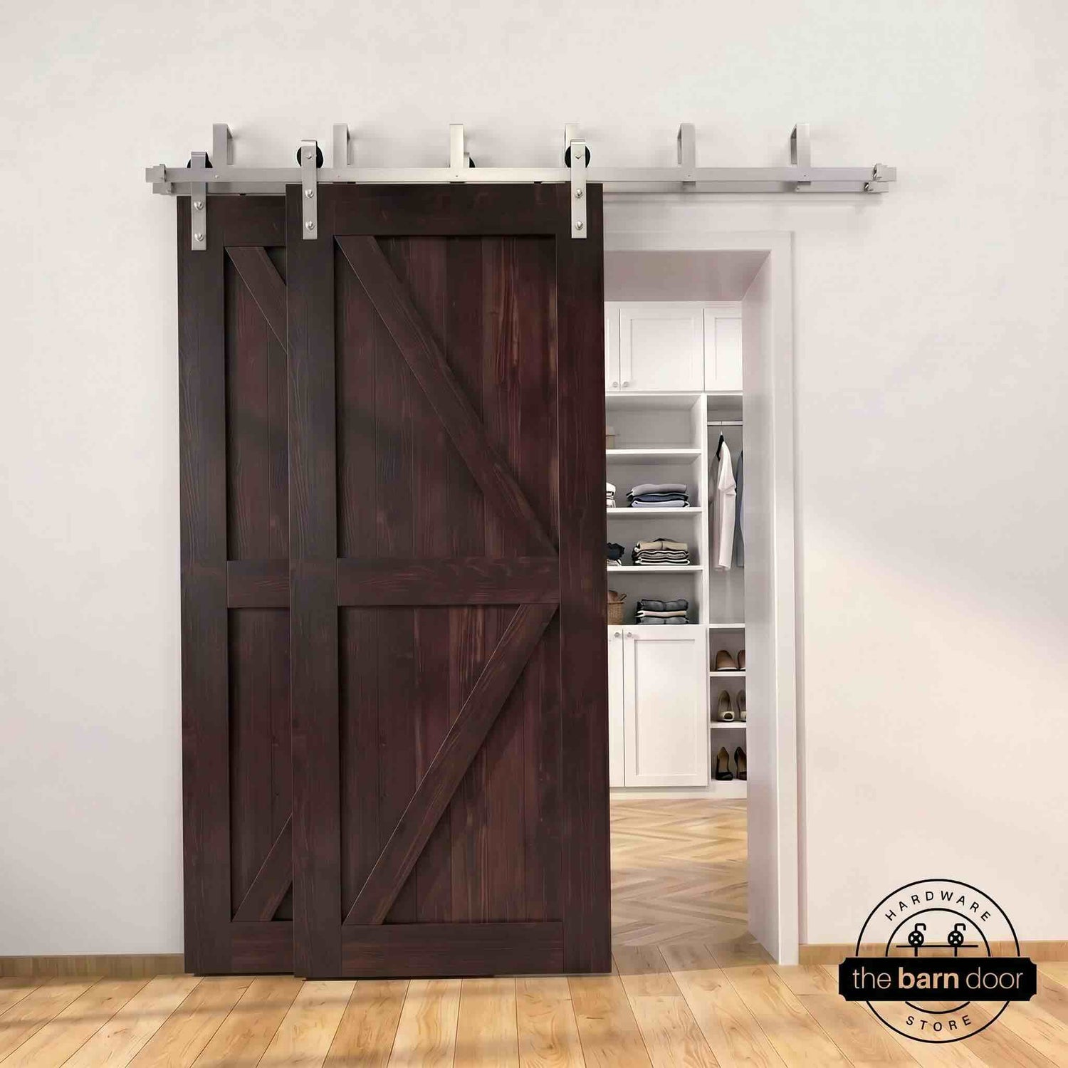Brushed nickel double bypass barn door kit with two red mahogany K-brace doors mounted on a dual-rail track, partially open to reveal a white built-in closet with folded clothing, hanging garments, and shoe shelving, set against a white wall with light herringbone and oak plank flooring.