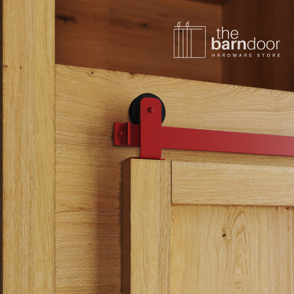 A close-up photograph of a sliding mini barn door on a natural oak cabinet, featuring red top-mount straight hardware.
