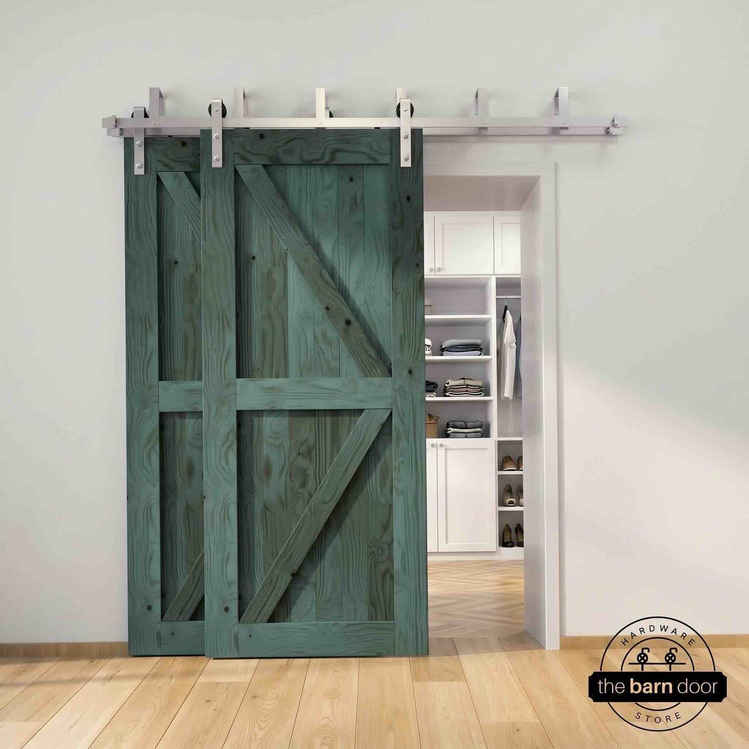 Brushed nickel double bypass barn door kit with two royal pine K-brace doors mounted on a dual-rail track, partially open to reveal a white built-in closet with folded clothing, hanging garments, and shoe shelving, set against a white wall with light herringbone and oak plank flooring.