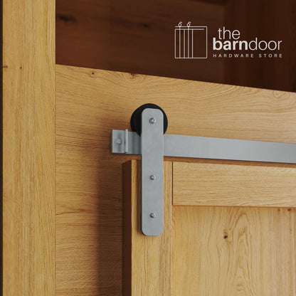 A close-up photograph of a natural oak cabinet with a sliding mini barn door, featuring a silver metallic straight-strap hanger and a silver metallic track system.