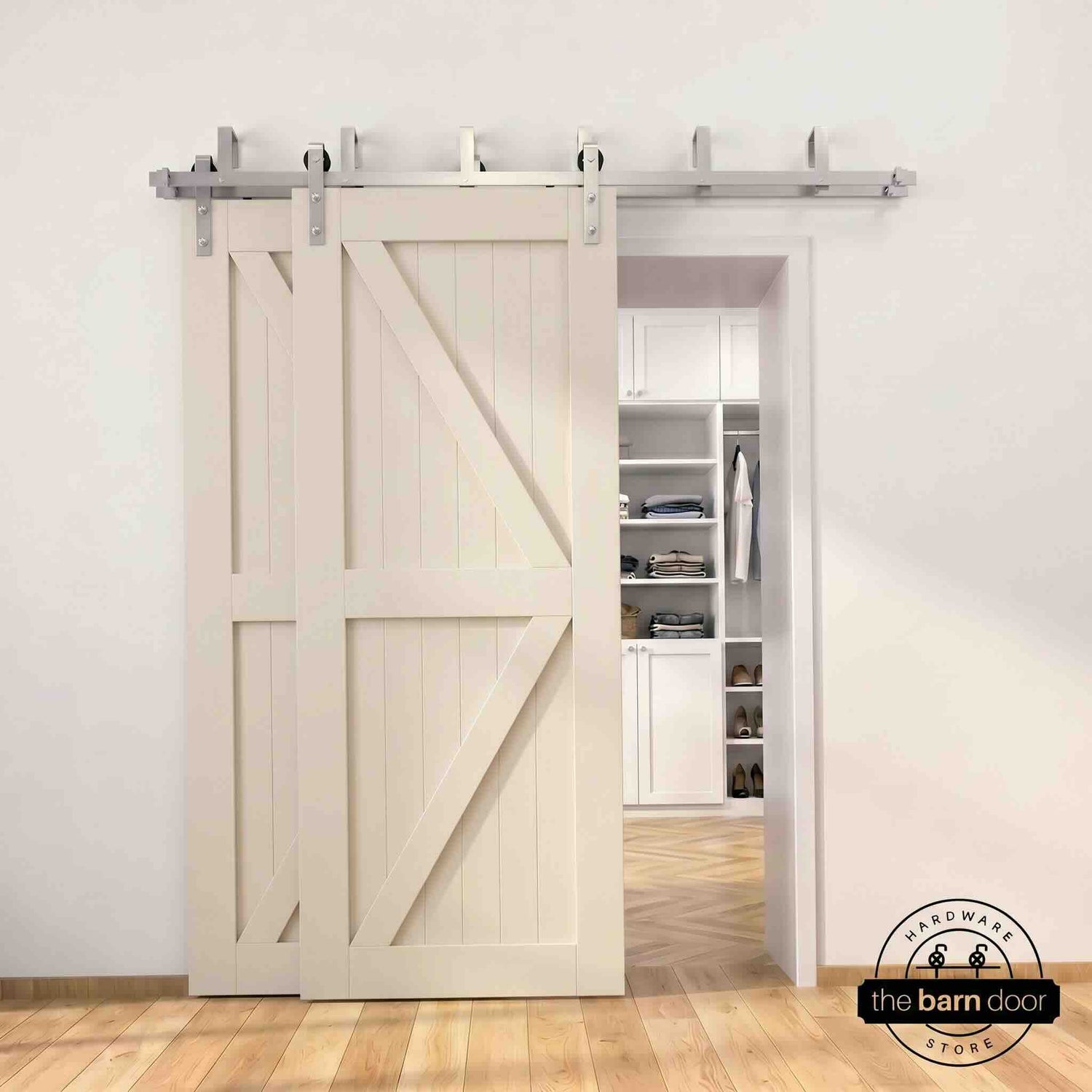 Brushed nickel double bypass barn door kit with two cream/off-white K-brace doors mounted on a dual-rail track, partially open to reveal a white built-in closet with folded clothing, hanging garments, and shoe shelving, set against a white wall with light herringbone and oak plank flooring.
