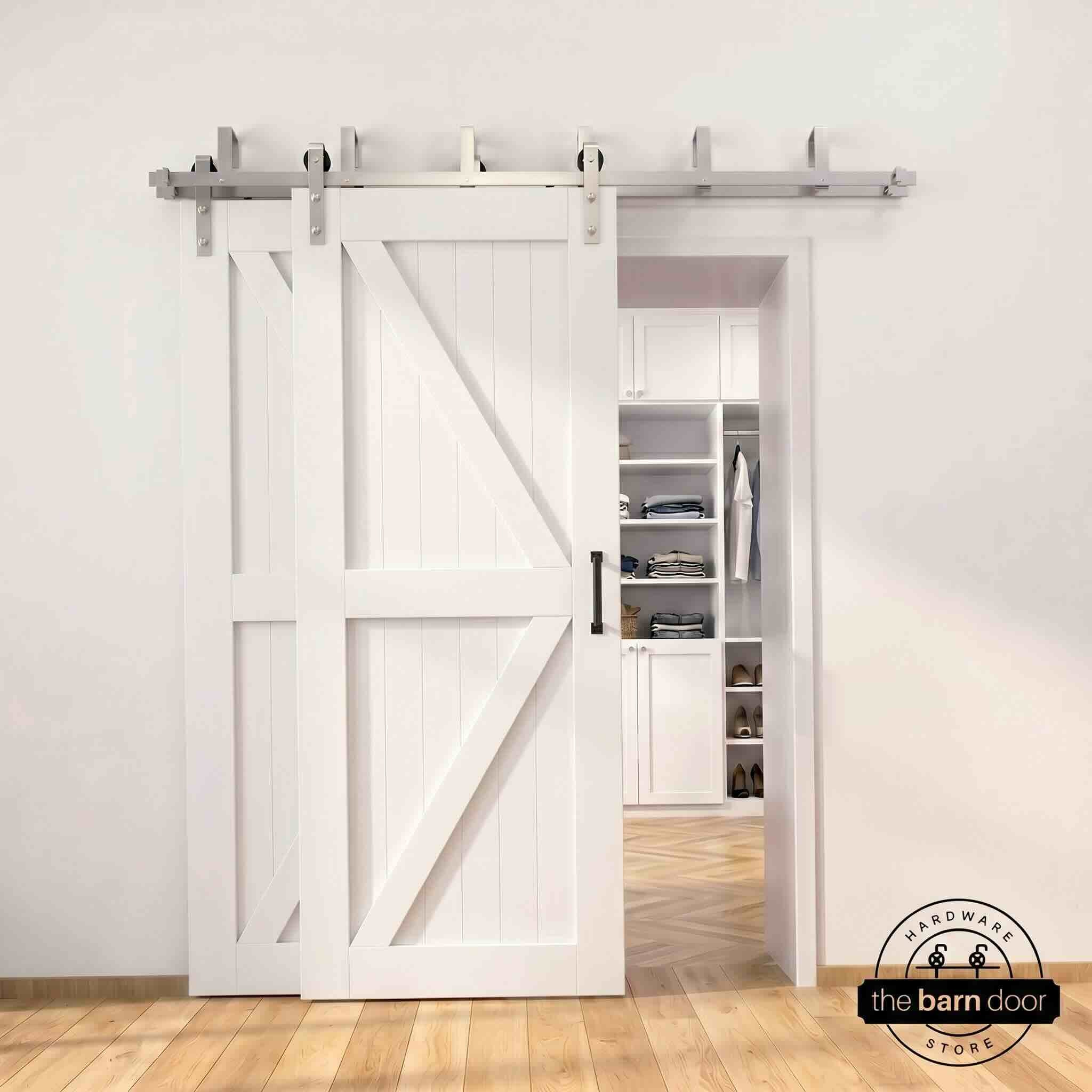Brushed nickel double bypass barn door kit with two white K-brace doors mounted on a dual-rail track, partially open to reveal a white built-in closet with folded clothing, hanging garments, and shoe shelving, set against a white wall with light herringbone and oak plank flooring.